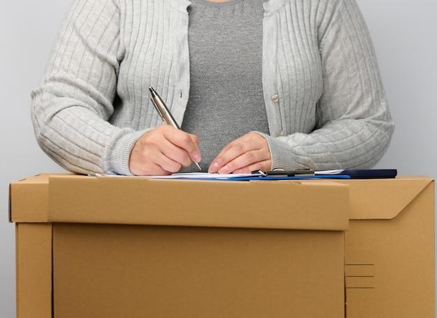 Une femme vêtue de gris se tient près d’une boîte brune et signe des documents avec un stylo. le concept de déplacement, d’envoi et de réception de marchandises. Licenciement